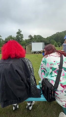 The video shows two individuals seated on a blue bench, facing away from the camera. The person on the left has vibrant red hair and wears a black denim jacket with sequined sleeves. The person on the right has dark, wavy hair and is wearing a loose-fitting shirt with a colorful floral pattern. They are both looking towards a white projection screen set up outdoors.

In the background, there are some tents or temporary structures, and beyond that, a wooded hillside under an overcast sky. The setting appears to be a grassy outdoor area, possibly a park or event space. A black bag with an orange accent is slung across the shoulder of the person on the right.

The activity appears to be an outdoor screening or presentation. The overall mood is relaxed, with the individuals seemingly attentive to the screen. The time of day seems to be daytime, given the ambient light, and the cloudy weather suggests a possibly cool or damp atmosphere. There are no visible signs with text in the provided frames. The location context places this scene in Fokino, Russia.