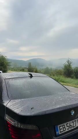 The video begins with a close-up of the rear of a black BMW sedan. The car's back window is wet with raindrops, and the license plate is partially visible, showing "EB 5627 BH." The car is parked on the side of a paved road. The view then pans to the right, revealing a misty, hilly landscape covered in lush green trees and vegetation. The sky is overcast with scattered clouds. The mist hangs low in the valleys, creating a serene and slightly ethereal atmosphere. The overall impression is one of a quiet, perhaps early morning, scene in a rural or mountainous region. There are no people visible in the video. The video seems to capture a moment of natural beauty and tranquility.