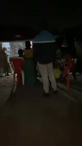 The video shows an outdoor night scene. Several people are gathered, some sitting on red plastic chairs and others standing. One person wearing a blue and white jacket and white pants is standing with their back to the camera. A person wearing a headscarf is near a brick wall. The camera then shifts focus to a person playing a video game on a smartphone, showing a football game screen. The lighting is dim, and the environment appears to be a gathering outside of a building or structure.
