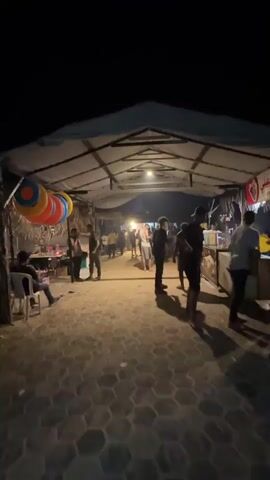 The video shows an outdoor marketplace at night, likely a street food or souvenir area. A group of people are visible walking around under a large, makeshift tent structure. In the foreground, a person in a black shirt and shorts walks towards the right. To the left, a person sits on a white chair. Hanging from the tent are several colorful inflatable rings, red, orange, yellow, and blue. Further down the path, various stalls are set up. One stall displays packaged goods on shelves. Another stall has a glass display case and signs with Arabic writing. A person in a black t-shirt is seen interacting with a stall. The ground is paved with interlocking stones. Several lights are suspended from the tent structure, illuminating the area. The overall atmosphere appears to be casual and lively.