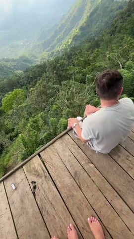 The video shows three people on a wooden platform overlooking a lush, mountainous landscape. The sky is overcast, with rays of sunlight breaking through the clouds. One person is lying down with their head propped up and wearing a red shirt and white socks. Another person is sitting up in a gray shirt. The person recording the video is standing and wearing a light blue shirt, white shorts, a woven Asian-style hat, and sunglasses. They have a tattoo on their arm. The camera angle is from the perspective of the standing person as they adjust their view to capture the scene.