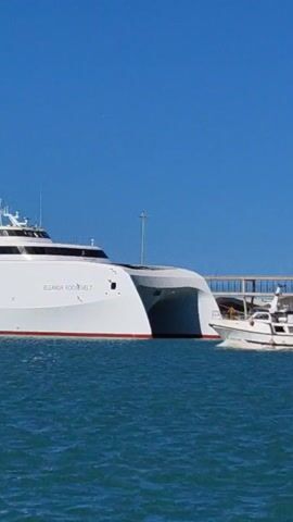 The video shows the ferry “Eleanor Roosevelt” berthed next to a pier on a sunny day. A small, white fishing boat passes in front of it. The sky is clear and blue, and the sea is a deep blue color.