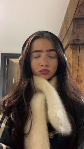 A young woman with long dark hair wearing headphones and a dark jacket with a cream-colored faux fur collar is looking directly at the camera. She adjusts the collar and runs her hands through her hair. The background includes a wooden beam and what appears to be a black-framed mirror or artwork. The lighting is bright and even. The woman's expression is neutral.  She has pink nail polish.