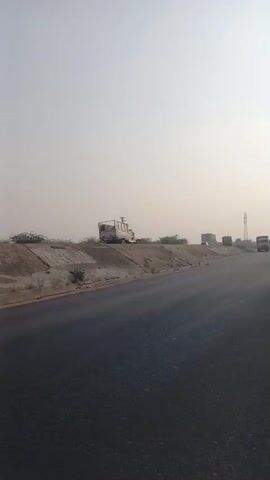 The video shows a view of a highway in what appears to be a rural area. A truck is driving towards the camera on the road. There is a small concrete barrier on the right side of the road, and beyond that there is a grassy embankment and some trees. In the distance, there are other trucks and what appears to be a cell tower. The sky is overcast and the light is hazy.