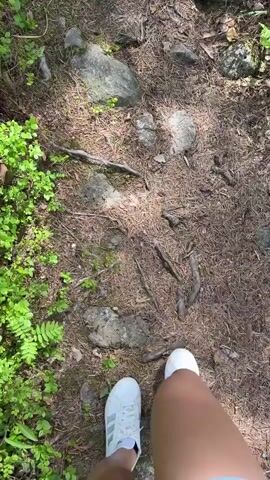 The video opens with a first-person perspective shot of feet in white sneakers walking on a forest path covered with pine needles, small rocks, and green undergrowth. The scene then transitions to a third-person view, showing a woman with blonde hair, wearing a black dress and black sandals, walking away from the camera on the same path. She carries a black shoulder bag. She walks into a sunlit clearing in a pine forest with tall trees and a blue sky overhead. The ground in the clearing consists of dirt, moss, and exposed rock.