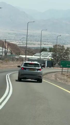 The video captures a scenic view of a road winding through a mountainous, arid landscape. The road is paved and marked with a yellow line. Several cars are visible on the road, including a gray SUV in the foreground, followed by a silver car. In the background, there are buildings and structures that appear to be part of a town or settlement, nestled in the valley below. Mountain ranges dominate the horizon, with a hazy, dusty atmosphere. A prominent green sign with white Hebrew text and an arrow pointing left is situated on the right side of the road. Streetlights are also visible along the roadside. The overall impression is a journey through a dry, mountainous region with signs of human habitation.