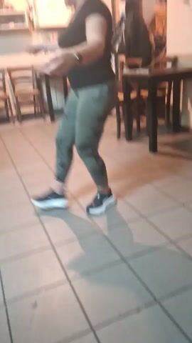 A woman in a black T-shirt and green pants is dancing in a restaurant. She has her hair pulled up in a messy bun, and is wearing a watch on her left wrist and a silver bracelet on her right. She’s wearing black and white tennis shoes. A woman stands behind her, clapping and smiling. There are several black tables and wooden chairs in the background. The floor is covered in gray tiles. There is a clock, an electrical outlet, and a fire extinguisher on the wall.