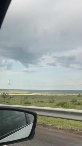 The video shows the view from a car window driving along a road. The landscape consists of a large open field with scattered vegetation under an overcast sky. Power lines are visible crossing the horizon. The side mirror of the car is visible in the lower left corner of the frame. The music is playing in the background.