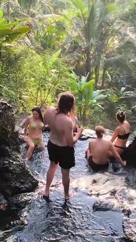 Four people are relaxing in a natural stream or waterfall setting. A woman in a yellow bikini sits on a rocky ledge to the left. Another woman, wearing a black bikini, sits on the opposite side. A man is sitting down in the water in the middle of the frame, while another man stands facing away from the camera. The area is lush with green foliage, including palm trees and banana plants. The water flows over rocks, creating a small pool. The scene is sunny.