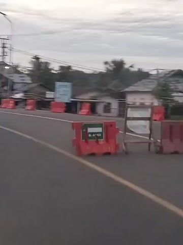 A medium-shot video captures the passing view from a moving vehicle. The scene shows a rural road with a two-way traffic lane on the left side of the frame and a roadside on the right. The road has a solid yellow line separating the two lanes and a solid white line marking the edge. A row of orange and white traffic barriers is visible on the roadside, with several small, rustic buildings behind them. Some of these buildings appear to be shops or residences. Overhead power lines crisscross the sky, supported by utility poles. A large green sign is partially visible on the roadside. The sky is overcast with a hint of morning light.