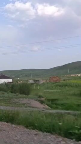 The video shows a moving view of a rural landscape with several houses scattered across rolling hills under a cloudy sky. The houses are simple, one-story structures with pitched roofs. There are also some utility poles and power lines visible. The foreground shows a dirt or gravel road and some vegetation, suggesting the video is being filmed from a vehicle.