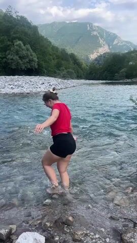 The video shows a woman standing in a shallow, clear river. She is facing away from the camera and towards the opposite bank, which is covered in white stones. Behind the river, there is a green forest and a mountain range under a partly cloudy sky. The woman has her hair in a bun and is wearing a red t-shirt and black shorts. She balances herself with outstretched arms, bends over to touch the water, and then stands up again.