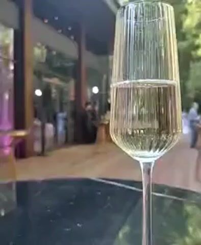 A champagne flute filled with a pale yellow liquid is in the foreground, positioned on a dark, reflective surface. The flute has vertical ridges. In the background, there is an outdoor event space with large glass windows, wooden flooring, and tables. Several people are visible in the background, some standing and interacting, while others appear to be mingling. The lighting suggests it is daytime or early evening, with some ambient artificial lighting also present. The overall scene depicts a social gathering or event.