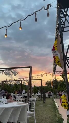 The video captures an outdoor event, possibly a wedding reception or party, set against a dramatic sunset. The sky is filled with colorful clouds in shades of orange, pink, and gray. In the foreground, a large metal structure with strings of lights and floral decorations is visible on the right side. To the left, tables draped with white tablecloths are set up on a lawn, with people seated and interacting. There are several trees and bushes surrounding the area, and in the distance, more lights suggest a larger gathering. A stage with a large screen displaying images is visible on the far right. The overall atmosphere appears festive and social.