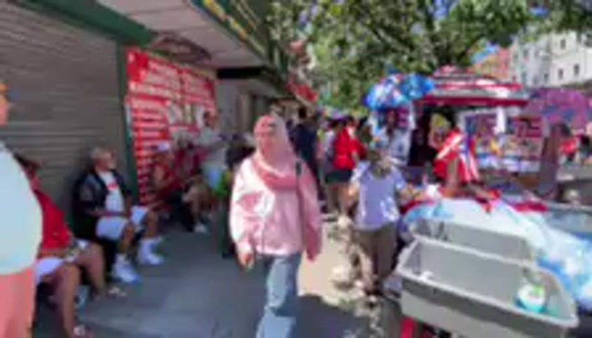 This media file depicts a crowded street scene in what appears to be a market or outdoor event in New York, United States. The time of day is clearly daytime, with bright sunlight casting shadows, suggesting fair weather.

The primary subjects are a large group of people of various ages and ethnicities. They are seen walking, standing, and interacting, creating a lively and busy atmosphere. Some individuals are wearing casual clothing, while others are dressed in more festive attire, including some with patriotic-themed clothing featuring red, white, and blue colors, possibly indicating a parade or celebration. One person in the foreground wears a pink hijab and a light-colored scarf. Other individuals are seen wearing hats, including a blue baseball cap and a fedora. There are also some objects visible, such as market stalls or vendor carts, decorated with colorful banners and fabric, some with red and white stripes. In the background, buildings and trees suggest an urban environment.

The activity appears to be a public gathering or street festival. People are moving through the street, and some are congregated around stalls or observing something. The overall mood seems to be energetic and celebratory, with people smiling and engaged.

There are some instances of visible text. On a stall to the left, red text on a white background is partially visible, but too blurred to decipher. Some banners also display text and graphics that are not clearly legible due to the resolution and motion of the video. The presence of what look like American flags suggests a patriotic event or holiday celebration.