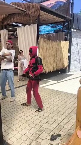 A person in a Deadpool costume is standing on a paved area with a thatched roof structure behind them. A man wearing a red bandana and a white t-shirt stands to the left of the Deadpool character, facing them. To the right of the Deadpool character, a woman in a patterned dress is standing and holding a phone. Other people are visible in the background, some sitting in cabana-like structures. The ground is paved with light-colored square tiles. The overall environment appears to be an outdoor recreational area or restaurant.