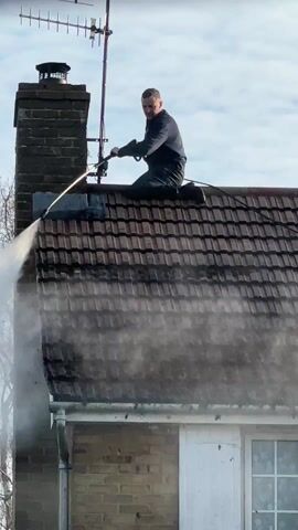 A man is kneeling on the tiled roof of a house, pressure washing it. He is wearing a dark long-sleeved shirt and dark trousers. His body is oriented towards the right side of the frame, and he holds a long-handled pressure washing wand with both hands, directing a powerful stream of water downwards and to the left. A significant amount of white mist is generated by the water hitting the roof tiles.

The roof is made of dark, possibly red or brown, tiles, and appears dirty or mossy in the unwashed areas. To the left of the man, a brick chimney rises from the roof, and behind it, a television antenna is mounted on a pole. The sky above is bright and shows some scattered white clouds. Below the roof, a light-colored brick wall of the house is visible, along with a white-framed window featuring multiple panes and white shutters. The man's left leg is bent with his knee on the roof, and his right foot is also planted on the roof. He appears focused on the task. No visible safety equipment like a harness is apparent. The overall scene suggests a bright, possibly sunny day.