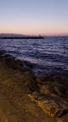 The video shows a view of the ocean at dusk. Waves are crashing against a rocky shoreline. In the distance, a lighthouse can be seen. The sky is a gradient of purple, pink, and blue. The camera pans to the right, revealing a paved walkway along the shore. The lighting suggests it is either early morning or late evening, with artificial lights illuminating the pathway. No people or animals are visible.