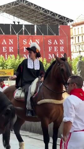 The video shows a parade or event featuring people on horseback. The individuals on the horses are dressed in traditional attire, including black jackets, white ruffled collars, and feathered hats. They are riding brown and black horses with ornate saddles. In the background, large red banners with white text that reads "SAN FER" are visible, suggesting the event is related to San Fermín. People in the foreground are watching the procession, some wearing white shirts and red sashes. The environment appears to be outdoors, possibly a street or plaza, with a stage or structure in the background.