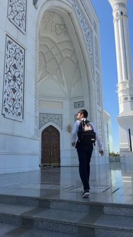 A young person with a backpack walks away from the camera towards a large ornate doorway. The person is wearing a white shirt and dark pants. The doorway is set within a grand white building with intricate carvings and architectural details. The ground is a reflective tiled surface, and the sky is a clear blue. The camera angle is low, looking up slightly at the building and the person.