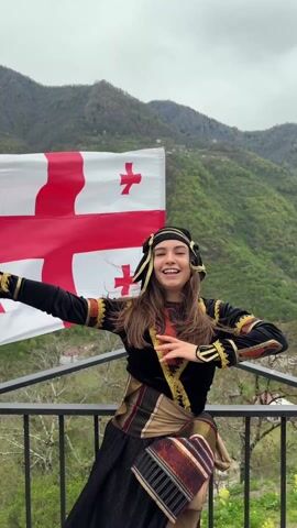 A young woman in traditional Georgian clothing stands on a balcony, laughing and posing in front of a large Georgian flag and a backdrop of green mountains. The woman has long brown hair and is wearing a black headdress, a black tunic with gold trim, and a patterned skirt with a woven belt. She is leaning against the balcony railing, her arms outstretched, and she is looking directly at the camera. The Georgian flag is waving behind her, and the mountains are visible in the distance. The sky is overcast.