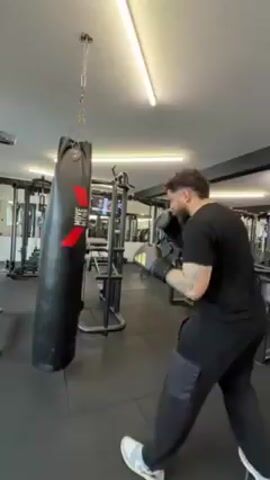 A man in black sweats and white sneakers punches a black heavy bag. He is wearing black boxing gloves or hand wraps. The bag is hanging in a gym, with various workout machines and weights visible in the background. The gym has a gray tile floor and bright overhead lighting. The camera angle changes drastically throughout the video, moving to follow the man’s movements and then losing sight of him as it focuses on the bag.