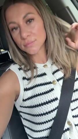 A woman with blonde hair is sitting in the driver's seat of a car. She is wearing a white and black striped sweater vest and a black seatbelt. Her right arm is raised, and she is touching her hair. Her left arm is resting on her lap. The background is blurred, but it appears to be an outdoor scene.