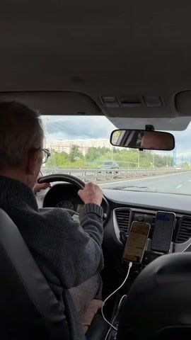 A gray-haired man is driving a car on a highway. He wears glasses and a gray sweater. His hands are visible on the steering wheel. On the dashboard, there are two smartphones mounted side by side, both displaying different applications. The car's interior is visible, including the rearview mirror reflecting the driver's face and the headrest of the passenger seat. Outside the car, trees and buildings are visible under a cloudy sky. Another car drives past in the opposite direction.