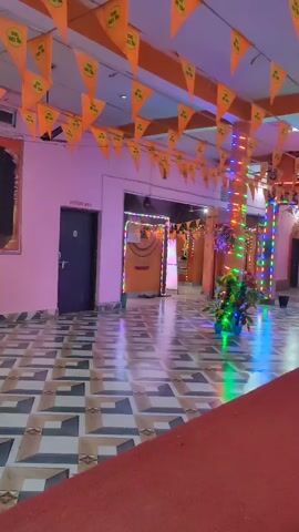The video shows the interior of a building decorated with orange triangular flags and colorful string lights. The floor has a patterned tile design. A man wearing a yellow t-shirt and blue shorts is sweeping the floor with a broom in front of a large framed picture depicting a religious scene. The picture shows three figures, presumably from Hindu mythology, with the central figure wearing an orange garment and a crown. There are potted plants with large leaves near a column decorated with string lights. The overall atmosphere appears to be festive or celebratory.