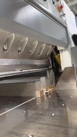 This video captures a close-up view of a large paper cutting machine in operation. The main subject is the cutting mechanism, a long, sharp blade descending to slice through a stack of paper. In the foreground, a metallic surface of the cutting table is visible, showing signs of use with scattered paper dust and circular indentations from fasteners. A small, partially cut object, possibly a sample or a test piece, sits on the table, showing the precision of the machine.

The machine itself is industrial-grade, with a substantial grey housing and visible internal components, including a yellow mechanical part. The cutting area is framed by a metal grate with a series of oval openings, likely for ventilation or debris removal. Above, a portion of a hand is visible, operating the machine, suggesting human involvement in the process.

The activity is clearly the precise cutting of paper. The descending blade and the stacks of paper indicate an ongoing operation, likely in a print shop, publishing house, or similar commercial setting. There are no explicit clues about the time of day or weather, but the indoor setting suggests controlled environmental conditions. No visible text is present on the machine or the materials being cut. The overall impression is one of industrial efficiency and precision. The location context of Ronnenberg, Germany, places this operation within a specific geographical setting, though the visual content itself is universally recognizable as a paper-cutting process.