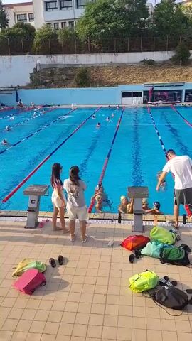 A swimming pool with multiple lanes and swimmers. On the pool deck, there are several people, some standing and observing, others preparing to enter the water. Bags and personal items are scattered on the ground. The scene appears to be a swimming practice or competition.