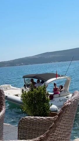 The video shows a white boat with a black canopy on blue water. There are at least four people on the boat. The boat has a flag on a pole at the back, which appears to be red and yellow. In the foreground, there is a partially visible wicker chair and a green bush. In the background, there is a coastline with hills and buildings. The sky is clear and blue. The boat has the word "Prince 30" written on the side.