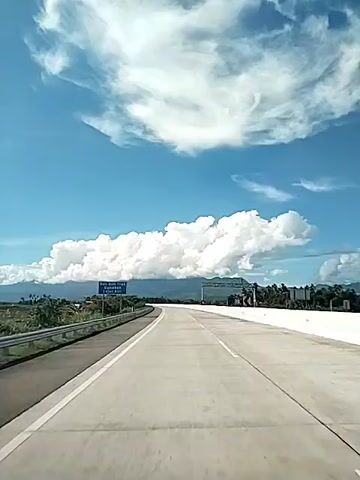 The video depicts a view from a vehicle moving along a wide, concrete highway on a bright, sunny day. The sky is a clear blue, filled with large, fluffy white clouds. On the left side of the road, there's a metal guardrail and a blue sign with white lettering. In the distance, mountains are visible, partially obscured by the clouds. Ahead, the highway curves slightly and a metal structure spans across the road. The scenery consists of greenery on either side of the road. The camera appears to be mounted inside the vehicle, providing a driver's perspective.
