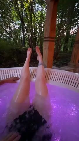 The video shows a first-person perspective from inside a hot tub at dusk or night, surrounded by trees and foliage. One person is relaxing in the hot tub with their legs extended upwards, visible above the bubbling, color-changing water. The water transitions through various vibrant colors, including pink, green, blue, and purple, illuminated by internal LED lights. Another person's hand is briefly visible resting on the edge of the hot tub, suggesting multiple individuals are present. Wooden support beams are visible to the right, indicating a structure built around the hot tub. The overall atmosphere is one of relaxation and enjoyment. A voice in Russian can be heard saying "Что ты делаешь?" (What are you doing?) at the beginning of the clip. The setting is an outdoor, possibly secluded, area, consistent with the provided context of Zakopane, Poland.