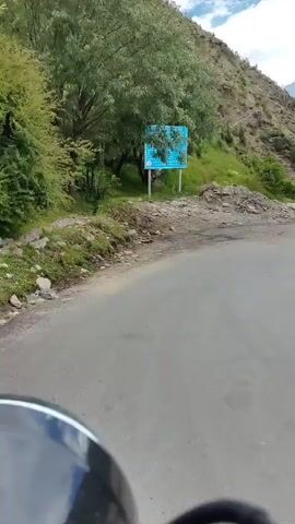 The video shows a view from the perspective of someone riding a motorcycle down a paved road that winds through a mountainous region. The road is on the right side of the frame, curving away from the viewer. To the left of the road, there is a steep, rocky mountainside covered in sparse vegetation. In the distance, a village can be seen nestled in a valley. The sky is cloudy. A signpost is visible on the left side of the road, near a tree. The motorcycle's headlight and part of its handlebar are visible in the foreground. The camera is moving, suggesting the motorcycle is in motion.