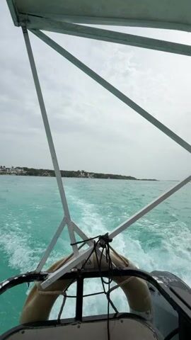 The video begins with a view from the stern of a boat, looking out at a turquoise body of water. White wakes trail behind the boat, indicating forward motion. In the distance, a lush green coastline is visible, dotted with white buildings and palm trees. The sky is overcast with gray clouds. As the video progresses, the boat continues to move, and the view of the coastline shifts slightly. A life preserver is visible hanging from the back of the boat. The camera pans slightly, revealing more of the vast expanse of water and the distant shore. The scene is serene and tranquil, suggesting a pleasant journey across the sea. The focus remains on the natural beauty of the tropical environment and the gentle movement of the boat.