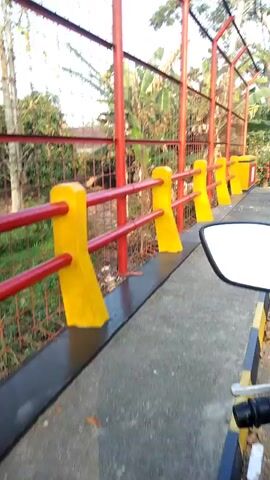 A person on a bicycle with a backpack rides past a fence with red poles and yellow accents. The fence is on the side of a paved path next to a road. Another person on a motorcycle drives past on the road. There are trees and a blue sky in the background.