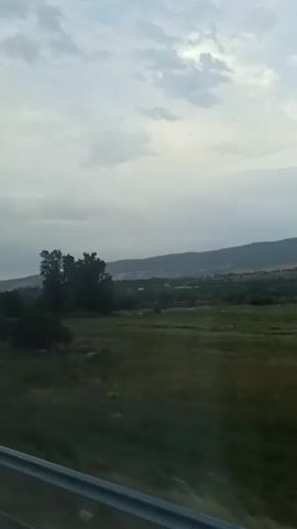 The video shows a view from a moving vehicle. The scene is a landscape with rolling hills covered in green vegetation. There are several trees scattered throughout the foreground and middle ground. The sky is overcast with some clouds visible. A gray railing is present in the bottom of the frame, indicating the perspective is from a window or a similar vantage point within a vehicle. The time stamps suggest a short duration, with the scene changing subtly as the vehicle moves.