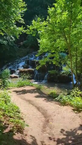 A dirt path leads to a multi-tiered waterfall in a lush green forest. Sunlight filters through the trees, casting dappled shadows on the path and surrounding vegetation. The water cascades over rocks and moss-covered formations, creating a serene and natural scene. The path is worn, suggesting it is a well-traveled route to the waterfall. Lush greenery, including ferns and trees with full canopies, dominates the landscape, creating a sense of tranquility and natural beauty.