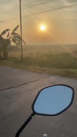 The video begins with a shot of a motorcycle's rearview mirror. The sun is rising in the background, casting a warm glow over a rural landscape. Banana trees are visible, along with a small house and a motorcycle parked nearby. As the video progresses, a person on a bicycle passes by. Further along the road, a pile of hay covered with a blue tarp is seen. Finally, a maroon-colored SUV is shown, with a person visible inside. The video appears to be filmed from a moving vehicle, likely a motorcycle, as it captures the surrounding environment from a first-person perspective.