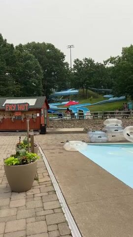 The video begins at a water park, showing a food stand labeled "Fry Hut" on the left. Beyond the food stand, several blue water slides wind down a grassy hill. In the foreground, a bright blue swimming pool stretches out, with numerous clear inflatable tubes stacked on the edge and floating in the water. A person is visible in the water slide area, sitting under a red umbrella. More people are scattered around the pool area, some sitting on lounge chairs and others in the water. The sky is overcast, and lush green trees surround the park. The video pans slightly to the right, revealing more of the lazy river and the surrounding landscape. Several pairs of flip-flops are scattered near the edge of the pool.