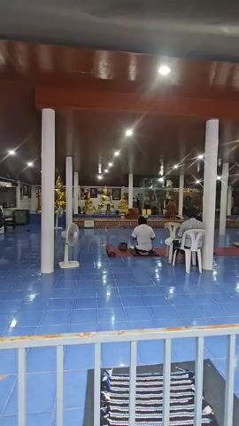 A medium shot captures the interior of a building, likely a temple or meditation hall, with bright blue tiled flooring and a warm wooden ceiling. Several large white pillars support the ceiling, and numerous recessed lights illuminate the space. In the background, a raised platform is adorned with many golden Buddha statues, suggesting a religious or spiritual setting.

The foreground reveals a serene scene with a few individuals engaged in prayer or meditation. Two people are visible in the center, sitting on yoga mats on the floor. One individual, seen from behind, wears a white shirt and is facing a small altar. Another person, also in white, sits in a similar posture to their left. To the right, a person in a striped shirt sits on a white plastic chair, also facing towards the altar. A floor fan stands near the left-side pillars.

The overall atmosphere is one of quiet contemplation and reverence. The lighting suggests it could be evening or an interior space with artificial illumination. The presence of Buddha statues and people meditating strongly indicates a Buddhist place of worship or a retreat. The location context provided places this scene in Ban Nai Rai, Thailand. No visible text is present in the footage.
