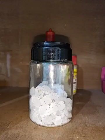 A clear glass jar with a black lid and a red dropper on top is filled with white, irregularly shaped crystals. Numerous small, dark ants are visible crawling on and around the crystals inside the jar. The jar is placed on a wooden surface, and the background appears to be a wooden shelf with other objects partially visible. The lighting is dim, casting shadows.