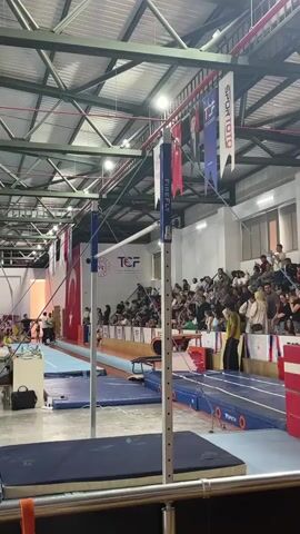 The video shows an indoor gymnastics competition. In the foreground, a women's uneven bars apparatus is visible, with blue mats on the floor around it. A gymnast in an orange and white leotard is performing on the bars, executing a routine. Spectators are seated in bleachers in the background, watching the competition. The gym has a high ceiling with exposed beams and lighting fixtures. Turkish flags and banners are displayed throughout the venue.