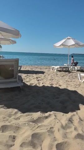 The video shows a sandy beach with lounge chairs and umbrellas. The ocean is visible in the background, with a yellow buoy floating in the water. There are a few people on the beach, including a woman standing near an umbrella and a man near a jet ski. The sky is clear and blue.