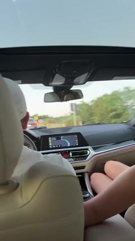 The video shows the interior of a car from the perspective of a passenger in the back seat. A person wearing a white cap is driving. A woman is in the passenger seat, her arm resting on the dashboard. The car has a large infotainment screen displaying a navigation map. The car is driving on a road with trees and other cars visible outside. The sun is visible through the sunroof.