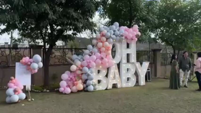 The video shows a baby shower setup on a grassy lawn. A large "OH BABY" sign, illuminated with lights, is decorated with pink, blue, and gray balloons. To the left of the sign, there is a smaller balloon arrangement and a welcome sign. Several people are standing in front of the display, including a woman in a long green dress, and a man taking photos. The background includes trees, a fence, and a building.