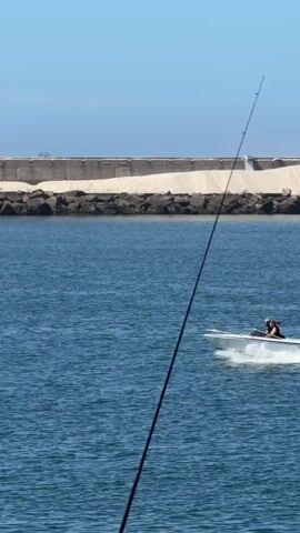 A white motorboat with two people aboard travels across a body of blue water. The boat is creating a white wake. In the background, a concrete seawall runs horizontally, with sand and rocks behind it. Above the seawall, a clear blue sky is visible.