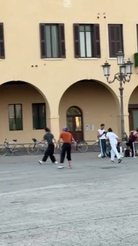 This media file captures a group of people engaged in what appears to be a dance or martial arts practice in an outdoor piazza. The setting is characterized by an Italian architectural style, featuring a large, pale yellow building with arched colonnades and dark-shuttered windows. A traditional-style street lamp stands prominently in the middle ground. Bicycles are parked along the colonnade, suggesting a public space frequented by cyclists.

The subjects are a diverse group, with at least five individuals visible and actively participating. Two individuals in the foreground are performing coordinated movements, one in a dark top and pants, the other in an orange top and dark pants. Further back, another individual in a white top and pants is engaged in similar activity, along with others who are less clearly defined in their actions. The atmosphere seems energetic and focused, with no obvious signs of distress or overt emotion.

The lighting suggests it is late afternoon or early evening, with soft, directional light casting some shadows. The sky is not visible, so weather conditions cannot be determined. The overall impression is of a lively urban square where people are utilizing the open space for physical activity. No discernible text is visible in the media file. The scene evokes a sense of community and shared engagement in a cultural or physical practice within a historic European setting.