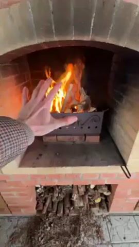 A close-up video shows a person's hand near a brick fireplace with a fire burning inside. The fireplace is made of red and light-colored bricks. Inside the firebox is a metal container filled with burning wood. The person moves their hand back and forth near the flames, seemingly testing the heat.  Stacked firewood is visible in a compartment beneath the firebox. The lighting is natural and suggests an outdoor setting or a well-lit room. The hand is wearing a patterned jacket.